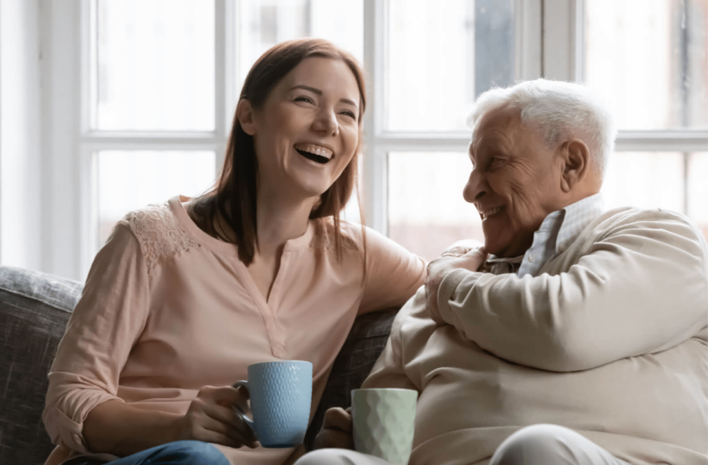 A caregiver laughing as she enjoys a cup of tea with a senior man. 