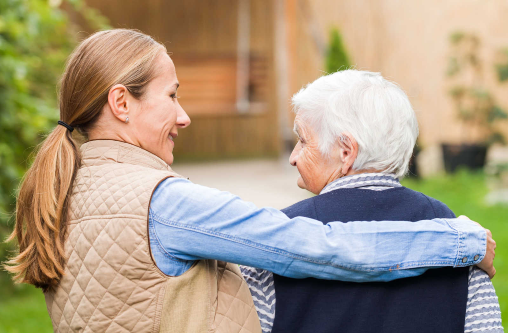 A female caregiver walking with an elderly woman with her arm around her shoulder.