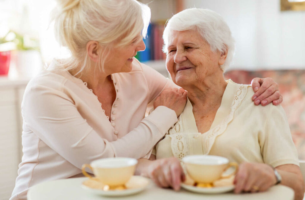 A senior woman and her daughter sitting on a couch smiling and talking to each other while having a cup of tea.
