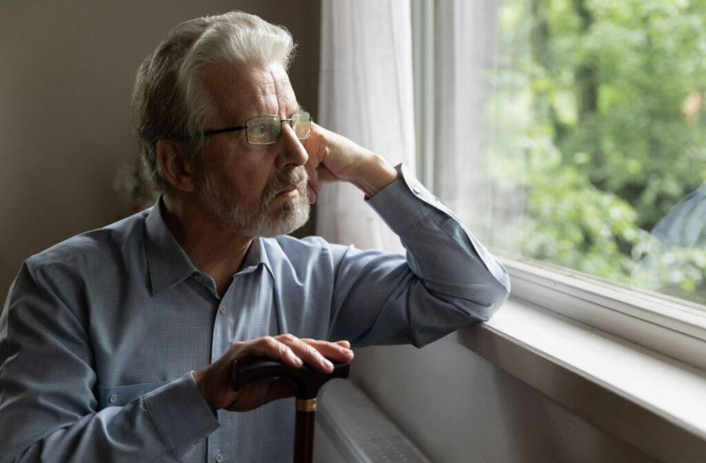 A senior man with glasses leaning on a windowsill and resting his right hand on the top of a cane while looking out the window with a serious expression