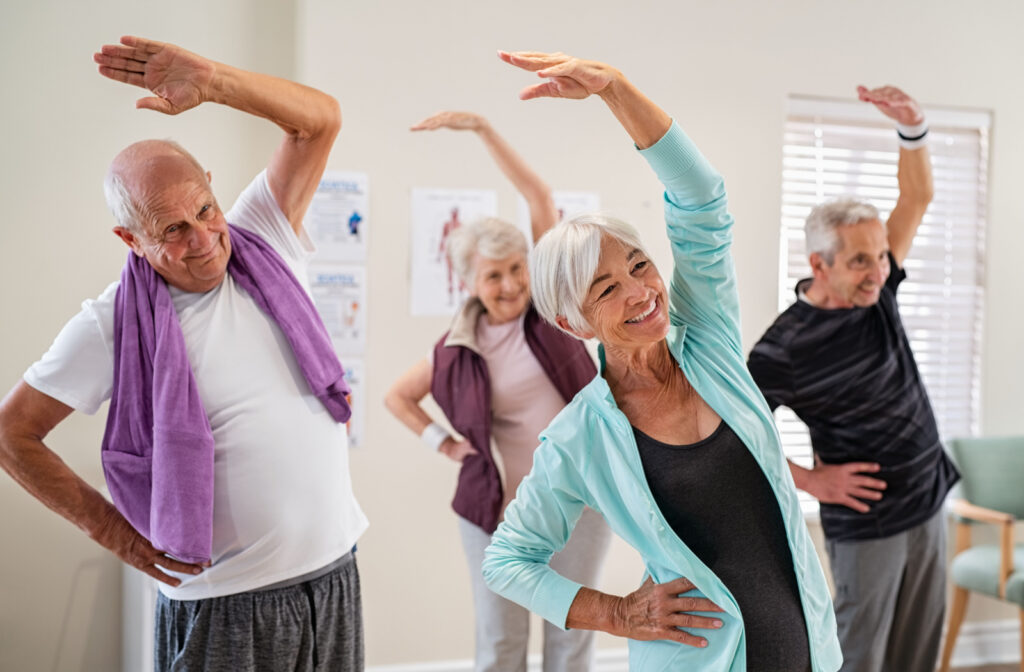 A group of seniors in an exercise class smiling and stretching with one hand over their heads