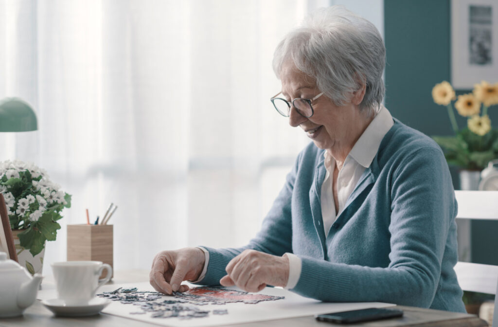 An older woman with glasses sitting at a table and doing a jigsaw puzzle with a cup of tea
