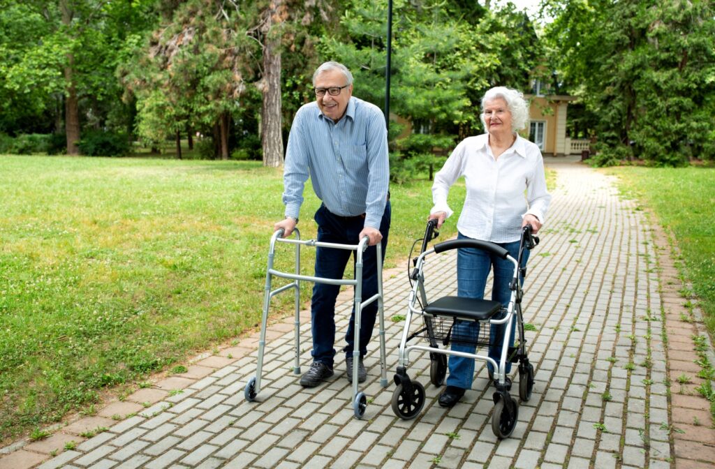 A happy senior couple walk through a park. One is using a two-wheeled walker while the other uses a rollator walker.