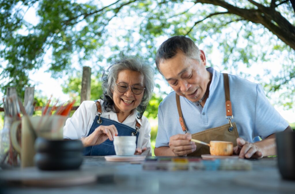 Two seniors laughing and conversing while painting homemade pottery at an outdoor table