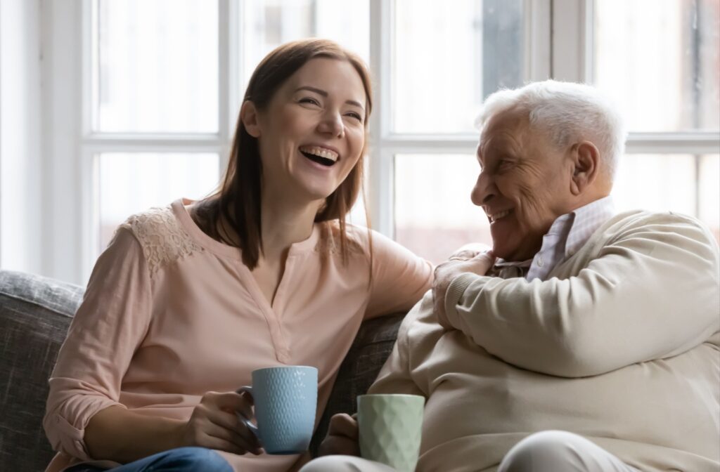 A senior and their adult child sitting on the couch laughing and enjoying a conversation together while they drinks a cup of tea