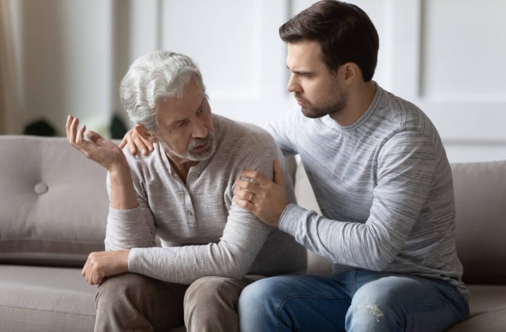 A stressed adult child holds their older parent on the couch during a difficult discussion about memory care