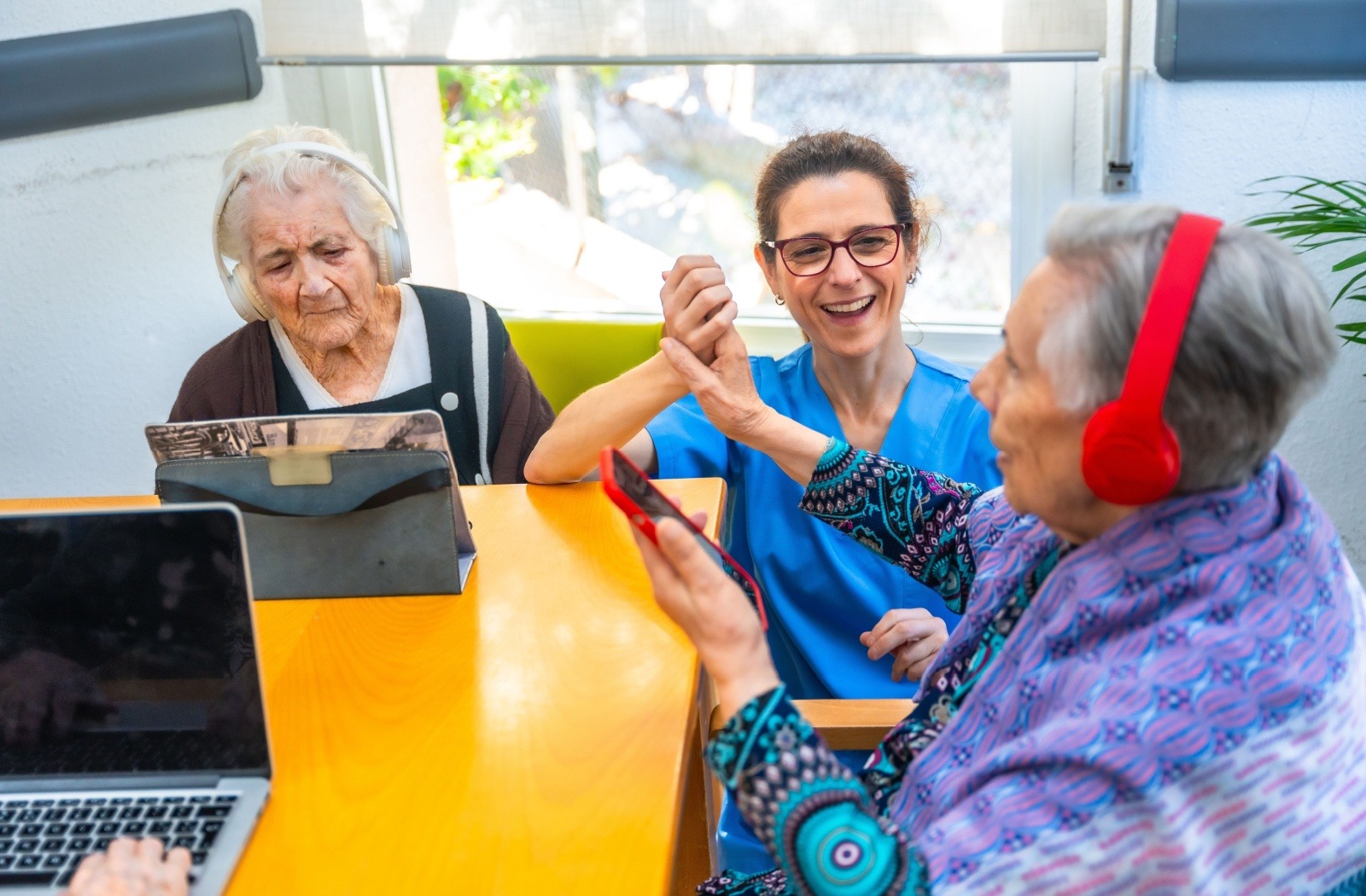 Older adults seated at a table listen to music using different computer devices and headphones while a smiling caregiver assists.
